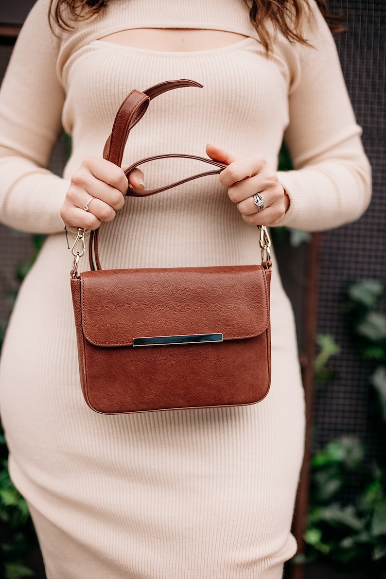 May include: A brown leather shoulder bag with a silver metal clasp. The bag has a long strap and is worn by a person in a white dress.