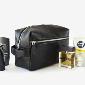 May include: A black leather toiletry bag with a silver zipper, handle, and various grooming products. The products include a bottle of cologne, face scrub, and other skincare items. The bag is rectangular and has a minimalist design.