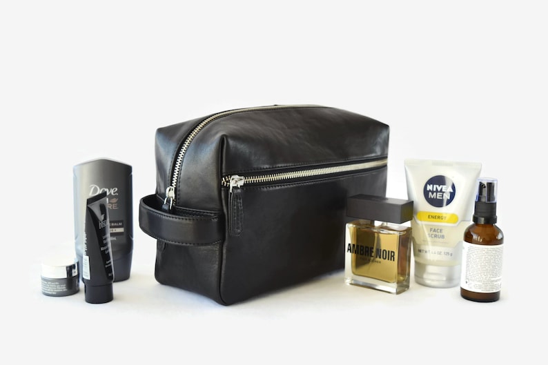 May include: A black leather toiletry bag with a silver zipper, handle, and various grooming products. The products include a bottle of cologne, face scrub, and other skincare items. The bag is rectangular and has a minimalist design.