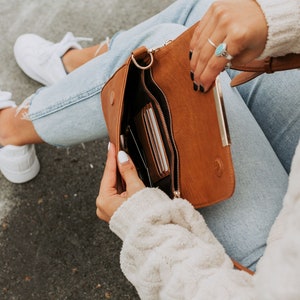May include: A brown leather crossbody bag with a gold zipper and a gold clasp. The bag is open and has several cards inside.