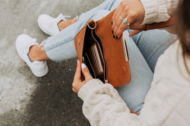 May include: A brown leather crossbody bag with a gold zipper and a gold clasp. The bag is open and has several cards inside.