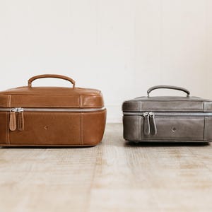 Two sizes of men’s leather toiletry bag shown side by side — large “Home” and compact “Travel” with top handles and dual zippers