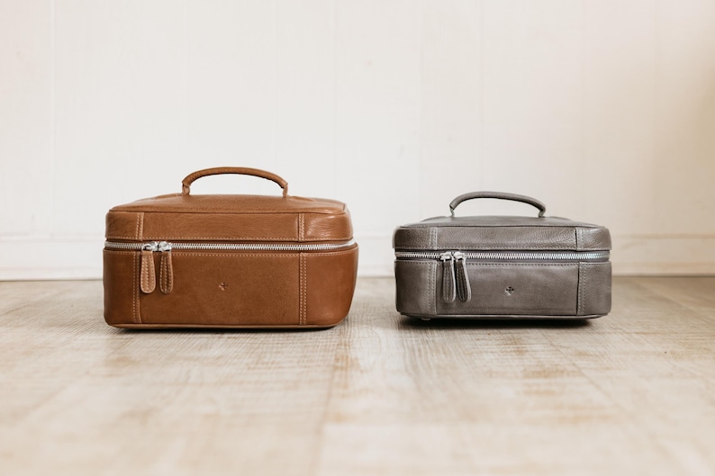 Two sizes of men’s leather toiletry bag shown side by side — large “Home” and compact “Travel” with top handles and dual zippers