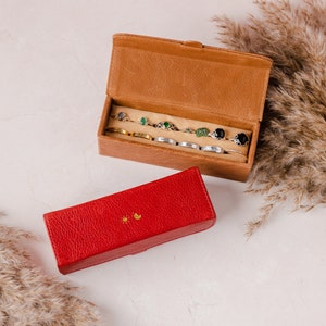 May include: A brown leather jewelry box with an open lid, displaying several rings with various gemstones. A red leather box with a sun and moon design is in the foreground. Dried ornamental grass adds a natural element to the composition.
