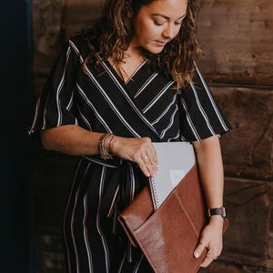 Coffee brown leather laptop sleeve carried under arm. Woman puts a notebook in the back pocket — slim work portfolio clutch for MacBook and notebooks.
