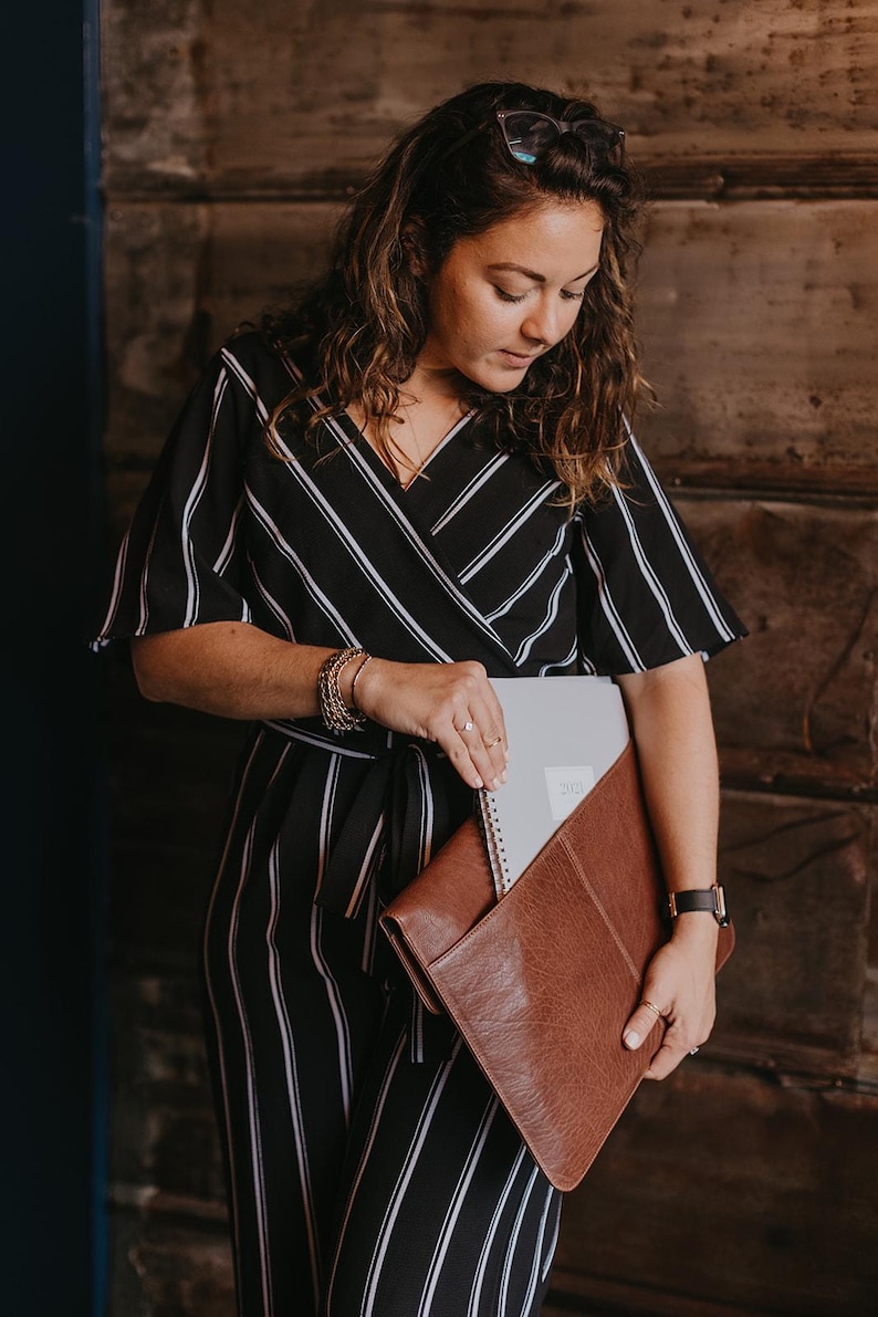 Coffee brown leather laptop sleeve carried under arm. Woman puts a notebook in the back pocket — slim work portfolio clutch for MacBook and notebooks.