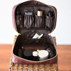 Può includere: Un beauty case da viaggio in pelle bordeaux con tiretti della cerniera dorati è aperto e pieno di vari prodotti per la cura della pelle, tra cui bottiglie, tubi e barattoli. La borsa è appoggiata su un tavolo in vimini.