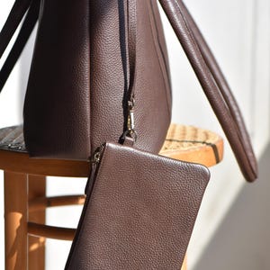 May include: A brown leather tote bag with matching wristlet. The tote bag has two handles and a long strap. The wristlet is rectangular with a zipper closure. Both items have a textured surface and are sitting on a wooden stool.