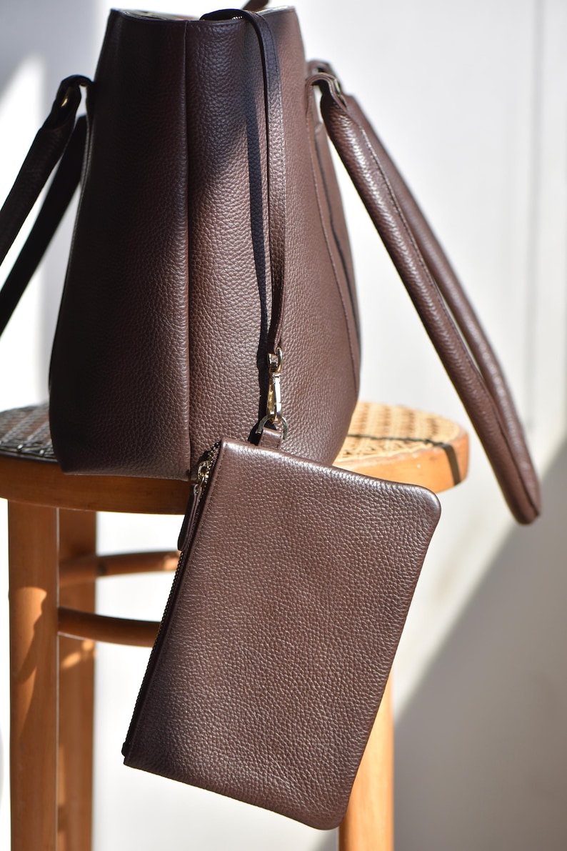 May include: A brown leather tote bag with matching wristlet. The tote bag has two handles and a long strap. The wristlet is rectangular with a zipper closure. Both items have a textured surface and are sitting on a wooden stool.