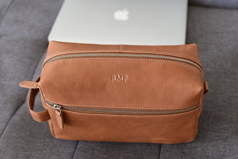 May include: A brown leather toiletry bag with two zippered compartments and a handle. The initials "RMF" are embossed on the front. The bag is sitting on a gray surface with a laptop in the background.
