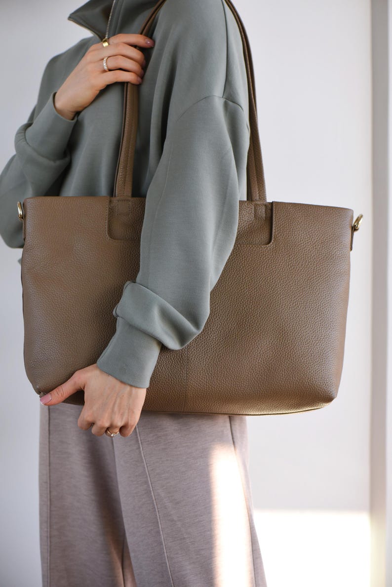 May include: A brown leather tote bag with a rectangular shape and two handles. The bag has a textured surface and a simple design. The bag is being held by a person wearing a green sweatshirt and tan pants.