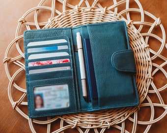 May include: Teal leather wallet with multiple card slots, a clear ID window, and a pen holder. The wallet is open and lying on a woven placemat.
