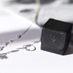 May include: A small black ring box with a heart embossed on the lid. The box is sitting on a white surface next to a sprig of lavender.