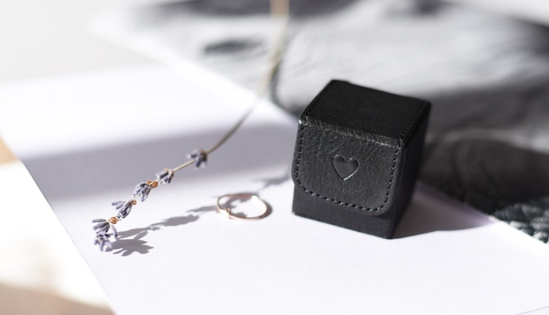 May include: A small black ring box with a heart embossed on the lid. The box is sitting on a white surface next to a sprig of lavender.
