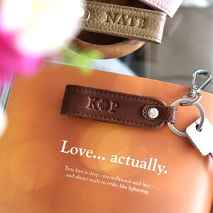 Op de afbeelding: Een bruin leren sleutelhanger met de initialen "KOP" erop, bevestigd aan een sleutel. De sleutelhanger ligt op een open boek met de tekst "Love... actually." Andere sleutelhangers met namen zijn zichtbaar.