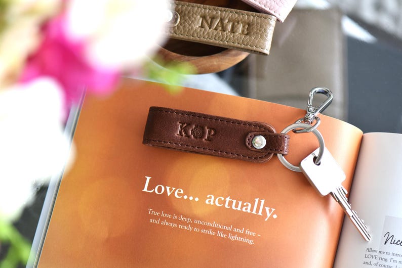 Op de afbeelding: Een bruin leren sleutelhanger met de initialen "KOP" erop, bevestigd aan een sleutel. De sleutelhanger ligt op een open boek met de tekst "Love... actually." Andere sleutelhangers met namen zijn zichtbaar.