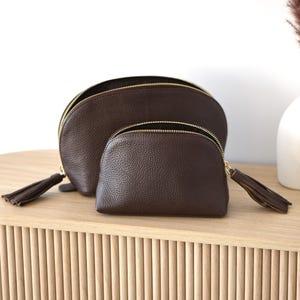 May include: Two brown leather cosmetic bags with gold zippers and tassel pulls. The bags are different sizes, with a larger, semi-circular bag behind a smaller, rounded bag. The bags are on a light wood surface.