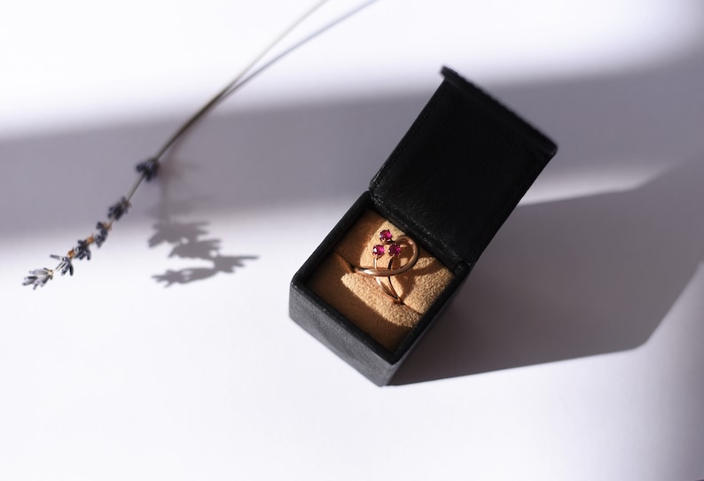 May include: A gold ring with three ruby stones set in a delicate, swirling design. The ring is displayed in a black velvet ring box.