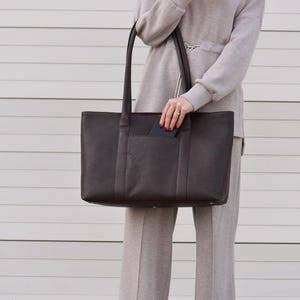 May include: A dark brown leather tote bag with long handles. The bag has a rectangular shape with a front pocket. The person is wearing a light brown turtleneck sweater and matching pants. The background is a white horizontal slatted wall.
