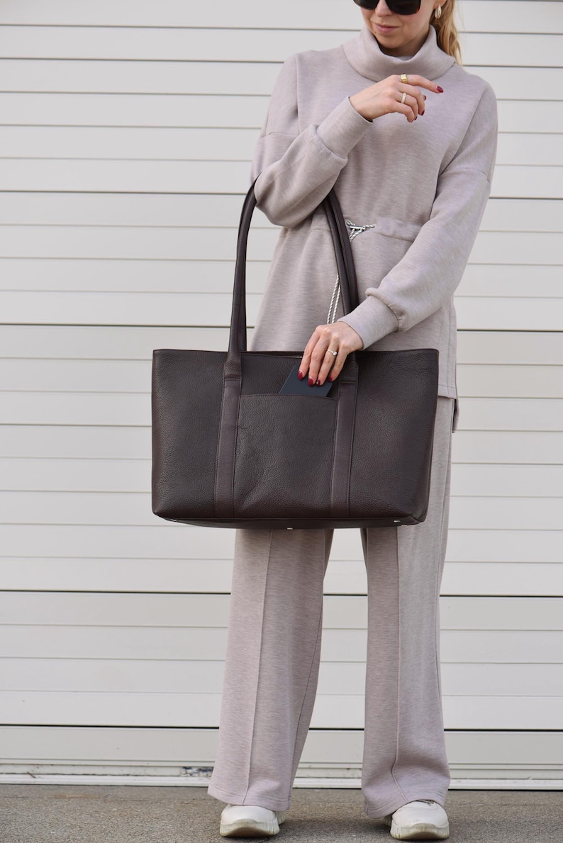 May include: A dark brown leather tote bag with long handles. The bag has a rectangular shape with a front pocket. The person is wearing a light brown turtleneck sweater and matching pants. The background is a white horizontal slatted wall.
