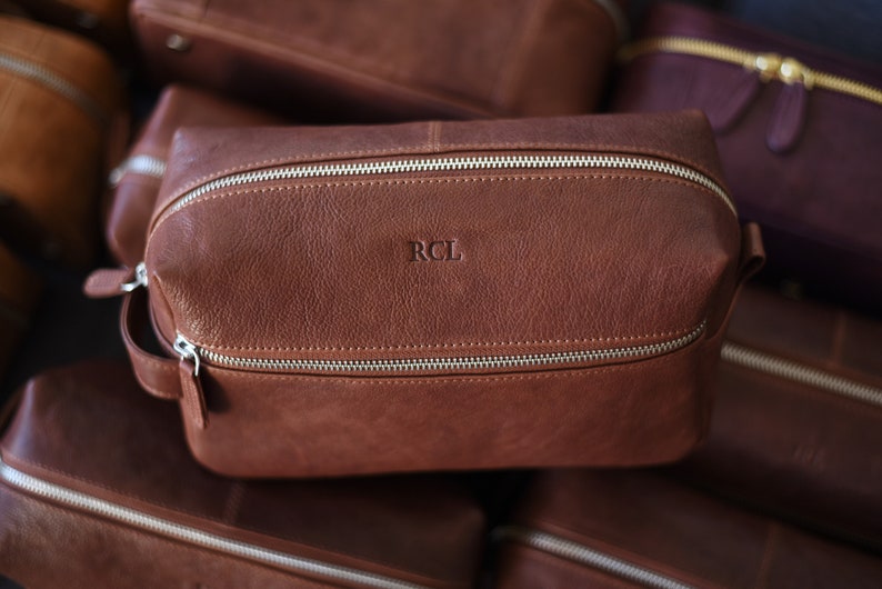 May include: A brown leather toiletry bag with silver zippers and a handle. The initials "RCL" are embossed on the front. Several similar bags are in the background.