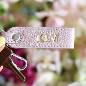 Op de afbeelding: Een lichtroze leren sleutelhanger met de initialen "KLV" in gouden letters. De sleutelhanger is bevestigd aan een zilveren sleutelring en een sleutel. De achtergrond is vervaagd met zachte roze en witte bloemen.