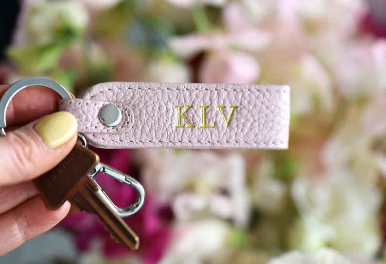 Op de afbeelding: Een lichtroze leren sleutelhanger met de initialen "KLV" in gouden letters. De sleutelhanger is bevestigd aan een zilveren sleutelring en een sleutel. De achtergrond is vervaagd met zachte roze en witte bloemen.