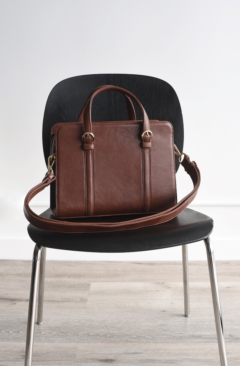 A brown full grain leather satchel bag with a top handle and a long adjustable shoulder strap. The bag has gold hardware and a zipper closure.