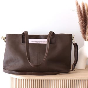 May include: A brown leather tote bag with a shoulder strap and handles. The bag has gold-colored hardware and a textured surface. A pink tag with text is inserted in the handles. The bag is on a wooden surface.
