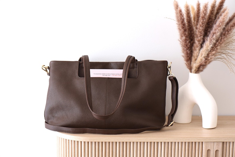 May include: A brown leather tote bag with a shoulder strap and handles. The bag has gold-colored hardware and a textured surface. A pink tag with text is inserted in the handles. The bag is on a wooden surface.