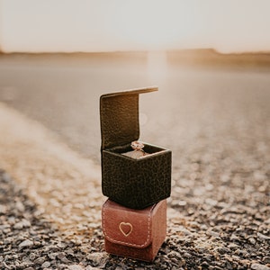 May include: A ring box with a diamond ring inside, sitting on top of a smaller heart-shaped box. The boxes are on a gravel road with a yellow line in the middle.