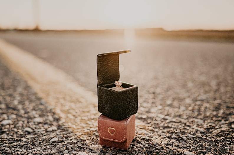 May include: A ring box with a diamond ring inside, sitting on top of a smaller heart-shaped box. The boxes are on a gravel road with a yellow line in the middle.