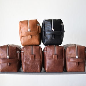 May include: A collection of leather toiletry bags in various shades of brown and black. Each bag features a rectangular shape, a silver zipper, and a small handle. The bags are stacked on top of each other, showcasing their design and texture.