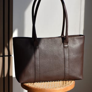 May include: A dark brown leather tote bag with two long handles. The bag has a rectangular shape and a textured surface. It is sitting on a small, round wooden stool with a woven seat.