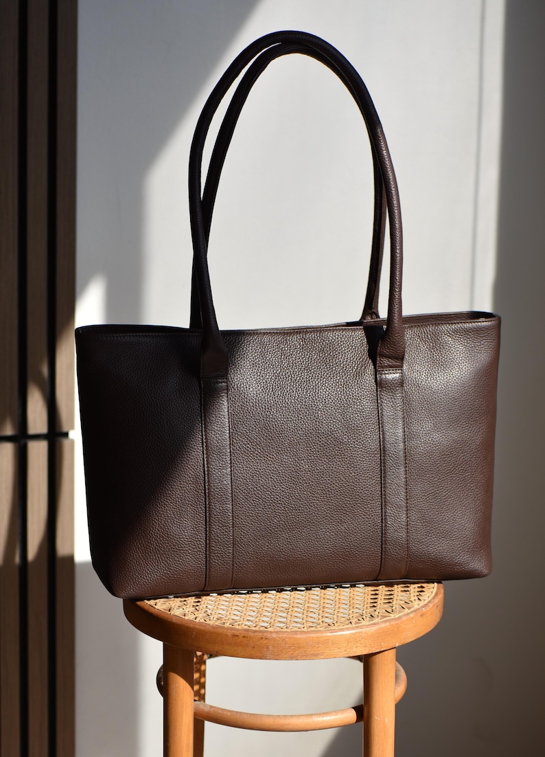 May include: A dark brown leather tote bag with two long handles. The bag has a rectangular shape and a textured surface. It is sitting on a small, round wooden stool with a woven seat.