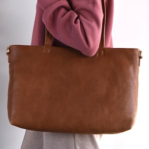May include: A large, rectangular brown leather tote bag with two handles. The bag is held by a person wearing a pink hoodie and gray pants. The bag has a simple, classic design.