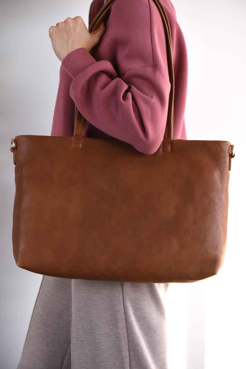 May include: A large, rectangular brown leather tote bag with two handles. The bag is held by a person wearing a pink hoodie and gray pants. The bag has a simple, classic design.