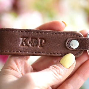 Op de afbeelding: Een bruin leren sleutelhanger met de initialen "KOP" en een rozenembleem in reli&euml;f op de voorkant. De sleutelhanger heeft een zilverkleurige metalen ring en een drukknoopsluiting. Het leer is gestikt met een contrasterende draad.
