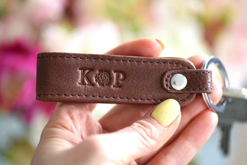 Op de afbeelding: Een bruin leren sleutelhanger met de initialen "KOP" en een rozenembleem in reli&euml;f op de voorkant. De sleutelhanger heeft een zilverkleurige metalen ring en een drukknoopsluiting. Het leer is gestikt met een contrasterende draad.