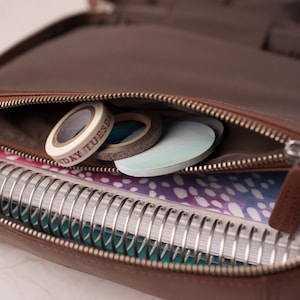 Puede incluir: Un estuche organizador con cremallera de aspecto cuero marr&oacute;n que contiene un cuaderno de espiral con una cubierta estampada en morado y blanco. El bolsillo frontal con cremallera contiene tres rollos de cinta decorativa, uno con el texto MONDAY TUESDAY.