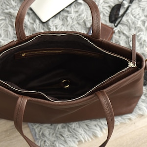 May include: Open view of a brown leather tote bag with two handles and a zipper closure. The interior is dark brown, and the bag appears to be empty. The zipper is gold-colored. The bag is sitting on a gray, fluffy surface.