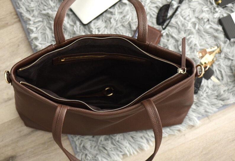 May include: Open view of a brown leather tote bag with two handles and a zipper closure. The interior is dark brown, and the bag appears to be empty. The zipper is gold-colored. The bag is sitting on a gray, fluffy surface.