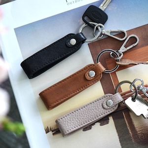 Op de afbeelding: Drie leren sleutelhangers in zwart, bruin en lichtgrijs. Elke sleutelhanger heeft een metalen ring en een druksluiting. Sleutels en een autosleutel zijn bevestigd.