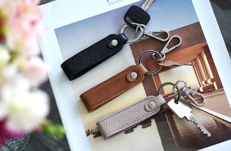 Op de afbeelding: Drie leren sleutelhangers in zwart, bruin en lichtgrijs. Elke sleutelhanger heeft een metalen ring en een druksluiting. Sleutels en een autosleutel zijn bevestigd.