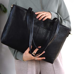 May include: A large black tote bag with long handles and a shoulder strap. The bag has a textured surface and a rectangular shape. The bag is held by a person wearing a green zip-up sweatshirt and tan pants.
