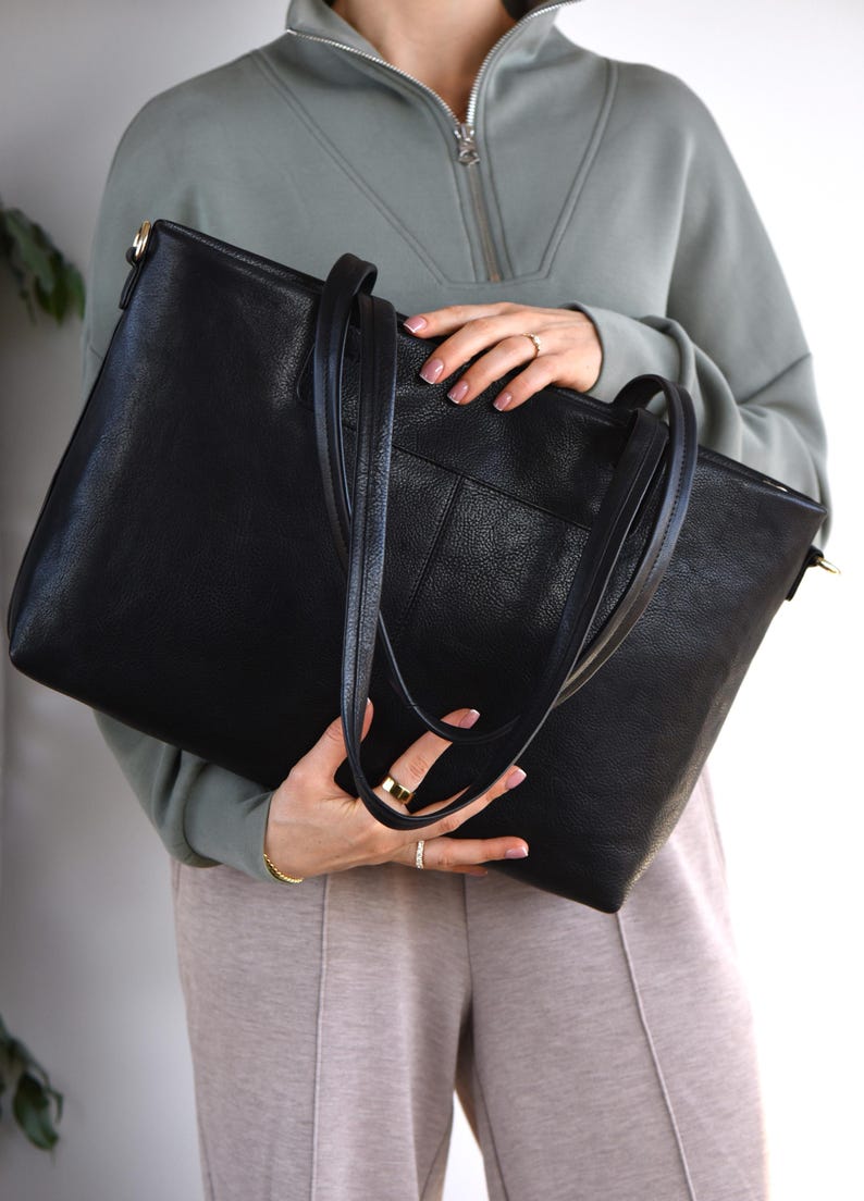 May include: A large black tote bag with long handles and a shoulder strap. The bag has a textured surface and a rectangular shape. The bag is held by a person wearing a green zip-up sweatshirt and tan pants.