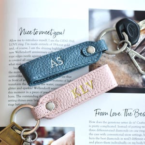 Op de afbeelding: Twee leren sleutelhangers, &eacute;&eacute;n in teal en &eacute;&eacute;n in roze, elk met initialen geborduurd in een contrasterende kleur. De teal sleutelhanger heeft "AS" in wit geborduurd, en de roze sleutelhanger heeft "KLV" in goud geborduurd. Sleutels zijn bevestigd.