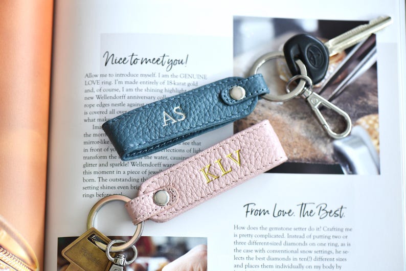 Op de afbeelding: Twee leren sleutelhangers, &eacute;&eacute;n in teal en &eacute;&eacute;n in roze, elk met initialen geborduurd in een contrasterende kleur. De teal sleutelhanger heeft "AS" in wit geborduurd, en de roze sleutelhanger heeft "KLV" in goud geborduurd. Sleutels zijn bevestigd.