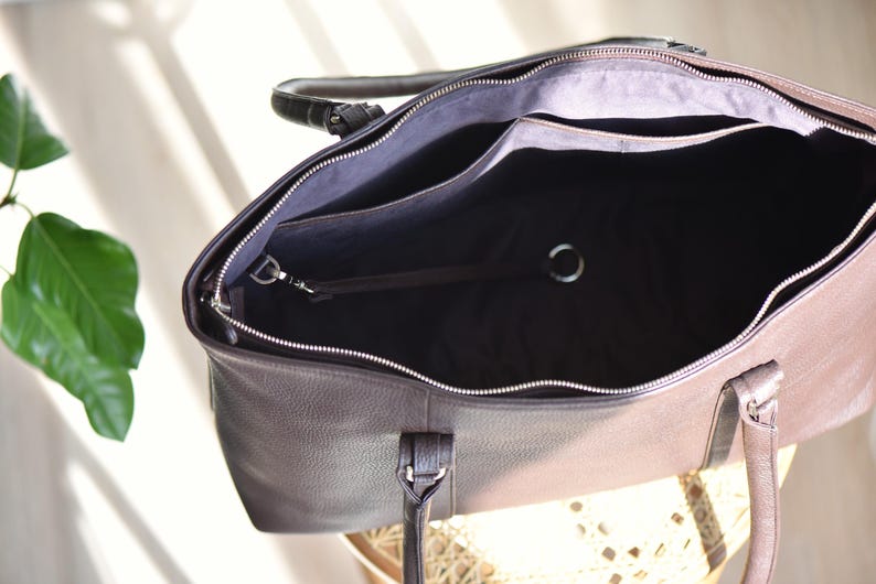 May include: Open view of a brown leather tote bag with a silver zipper and handles. The interior is black with pockets and a silver ring. The bag is sitting on a light-colored surface.