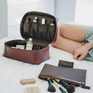 Two brown leather travel cosmetics bags with zippers, one open to reveal compartments and bottles, the other closed with makeup brushes inside. A small compact mirror and two makeup compacts are on a white surface.